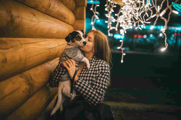 Eine Frau hält ihren kleinen Terrier vor einer Holzhütte im Arm und gibt ihm ein Küsschen.