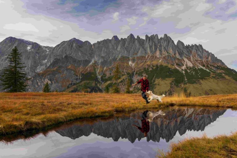 Ein Mann spielt vor traumhaftem Bergpanorama mit seinem Golden Retriever an einem kleinen See.